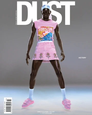 Dark-skinned male model posing in pink t-shirt, skirt, socks and sandals, wearing a durag and a necklace against a grey backdrop.