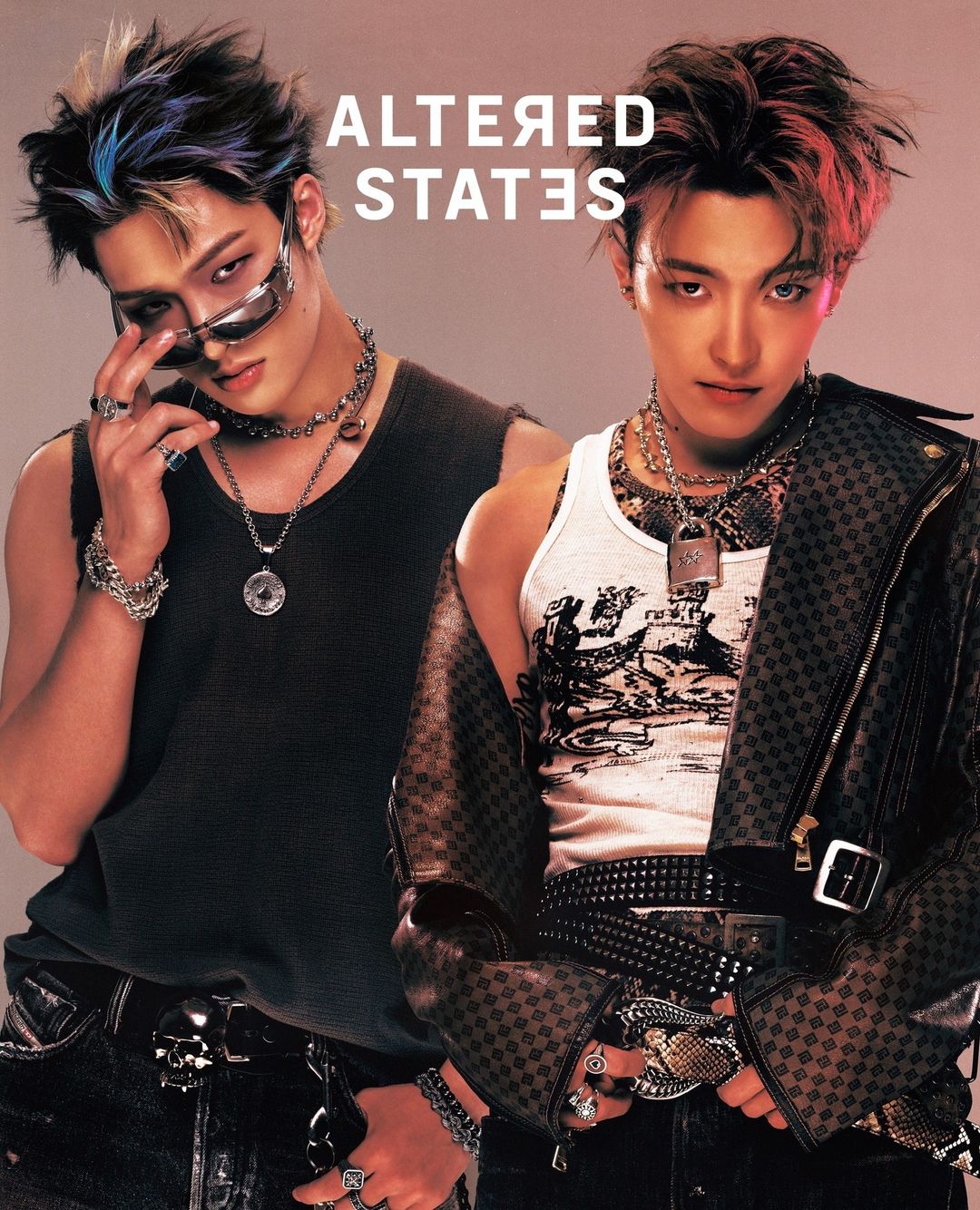 Two stylish young men pose indoors wearing fashionable casual clothing, accessories, and edgy hairstyles in front of a blurred background.