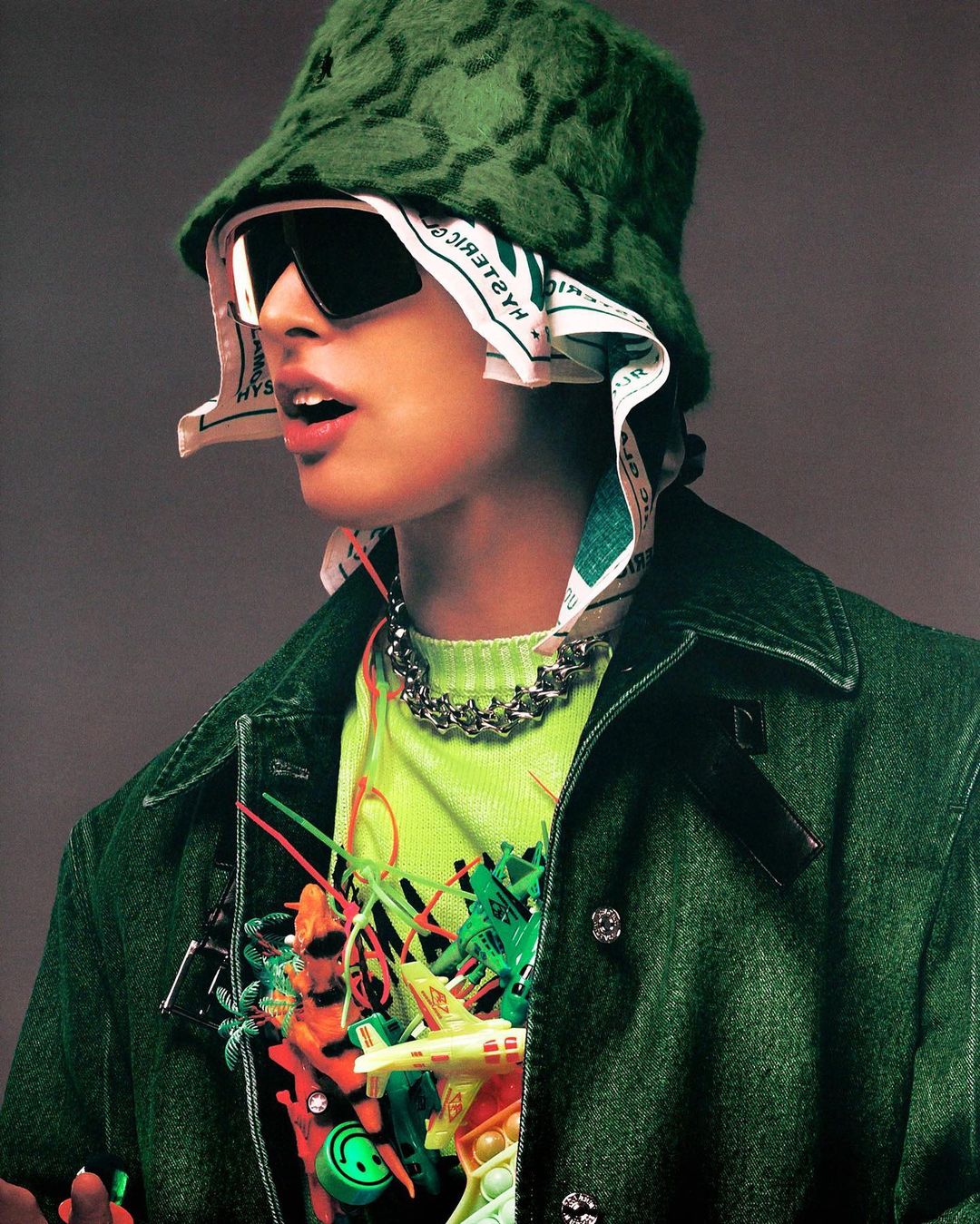 Fashionable person in a green bucket hat and denim jacket, accessorized with a statement chain and colorful toys pinned to the jacket.