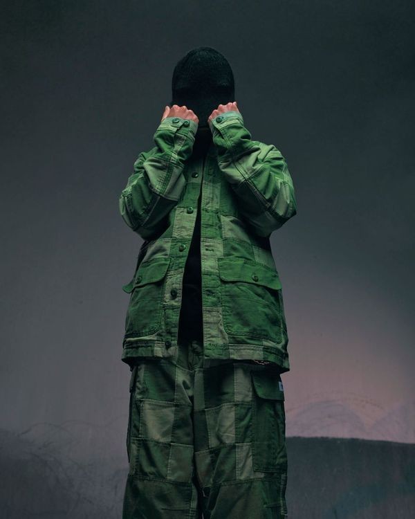 Person in green patchwork jacket and pants stands in front of a grey background, face obscured by hands and a dark beanie.