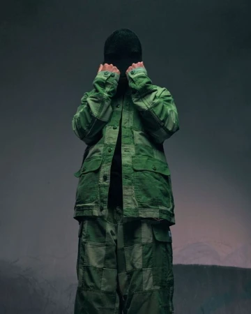 Person in green patchwork jacket and pants stands in front of a grey background, face obscured by hands and a dark beanie.