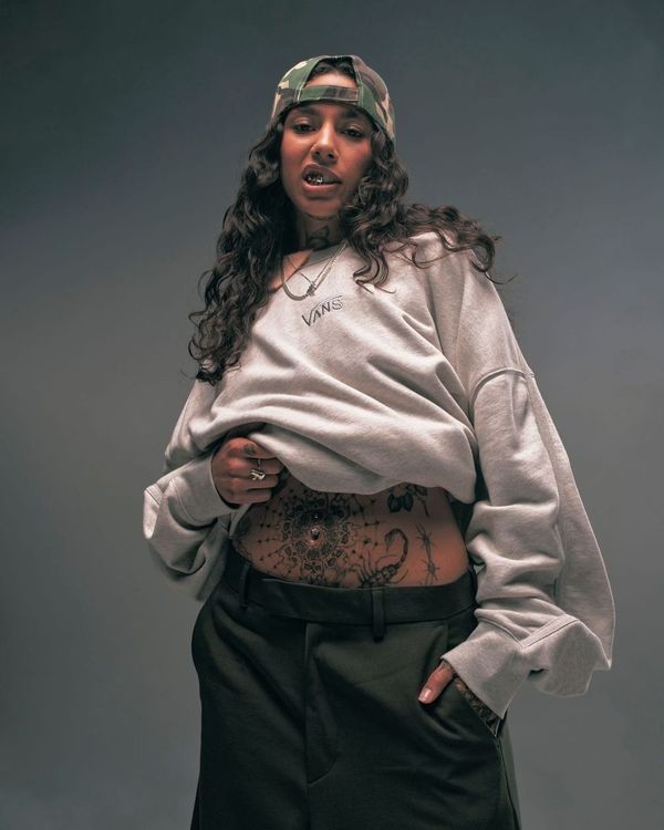Portrait of a woman wearing a grey sweatshirt, camo cap, and dark pants posing in front of a grey backdrop, showing midriff tattoo.
