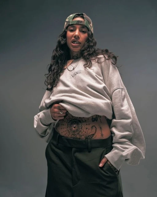 Portrait of a woman wearing a grey sweatshirt, camo cap, and dark pants posing in front of a grey backdrop, showing midriff tattoo.