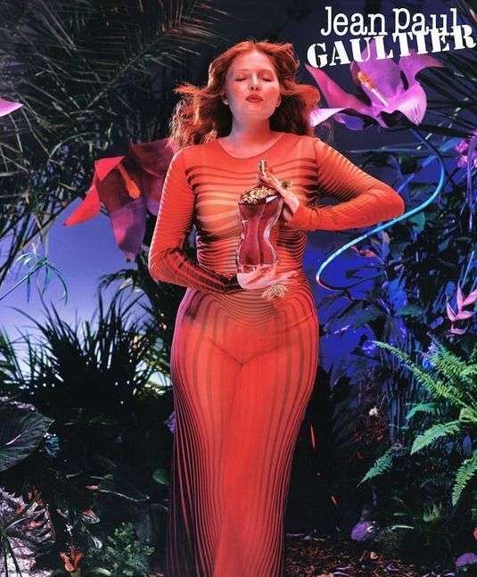 Adult female posing in a sheer orange dress, holding a perfume bottle in a lush botanical setting, with Jean Paul Gaultier text overlay.