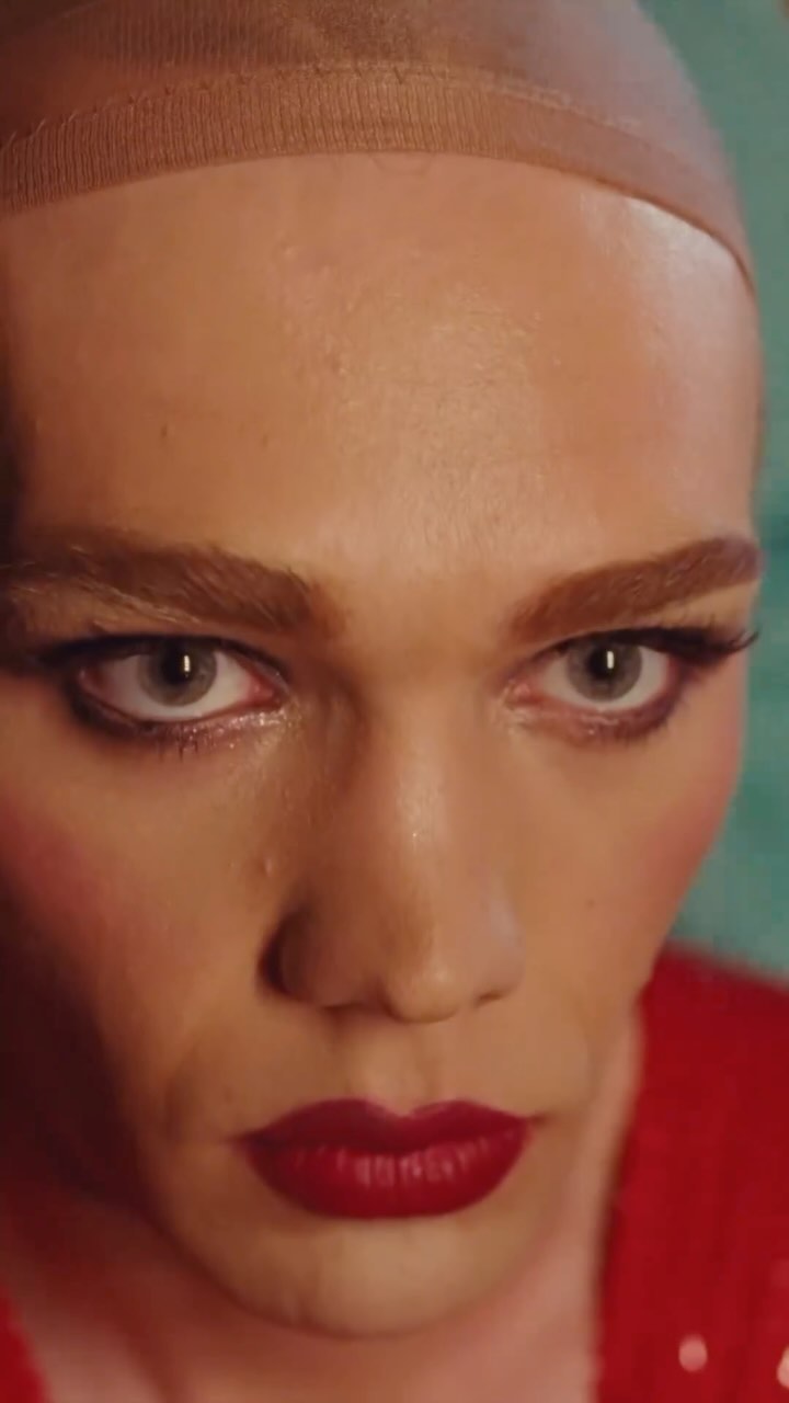 A close-up of a person wearing a skin-toned wig cap and red lipstick, looking directly at the camera with a serious expression.