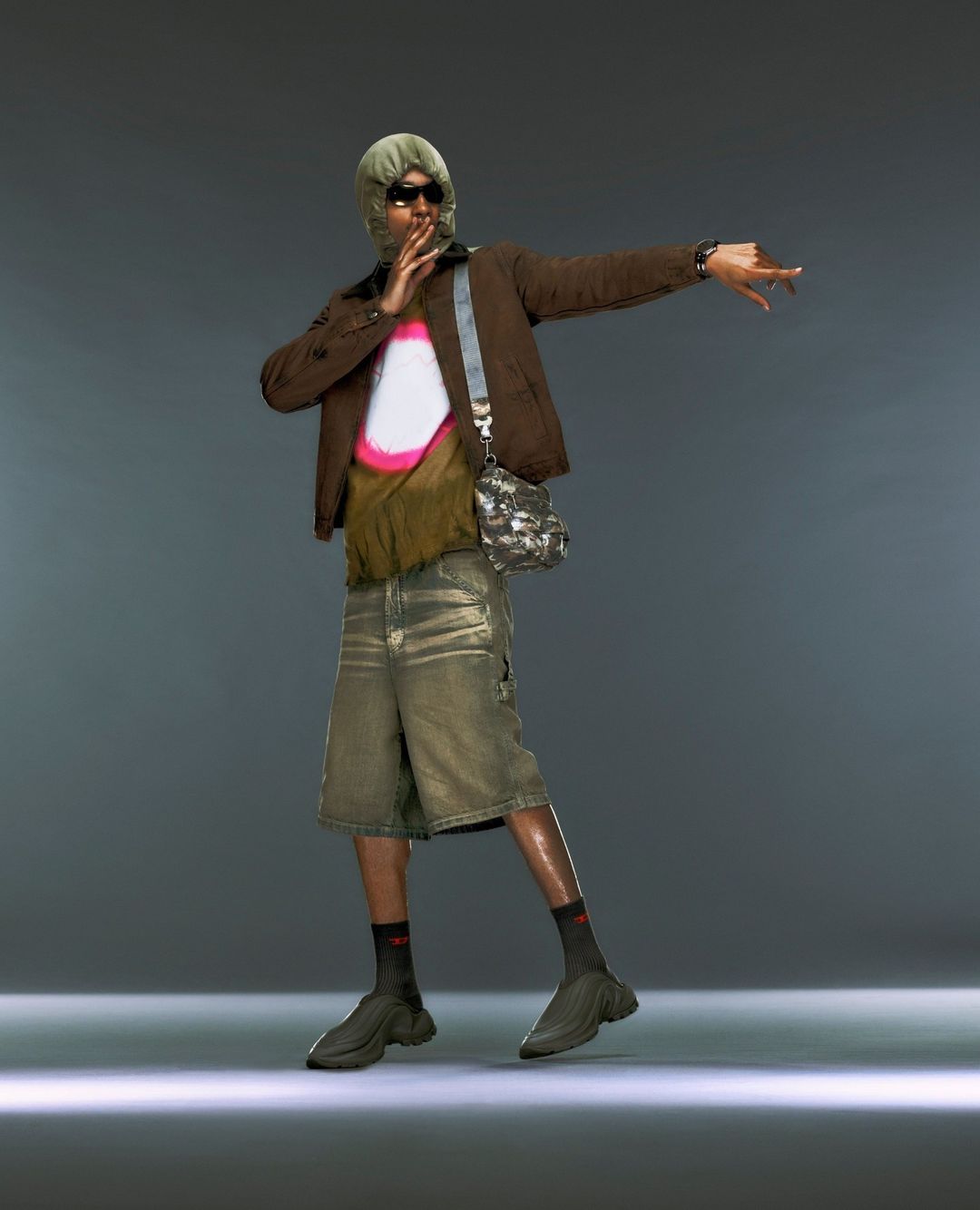 A person in a light green balaclava, brown jacket, graphic tee, and shorts poses in a studio with a cross body bag and athletic shoes.