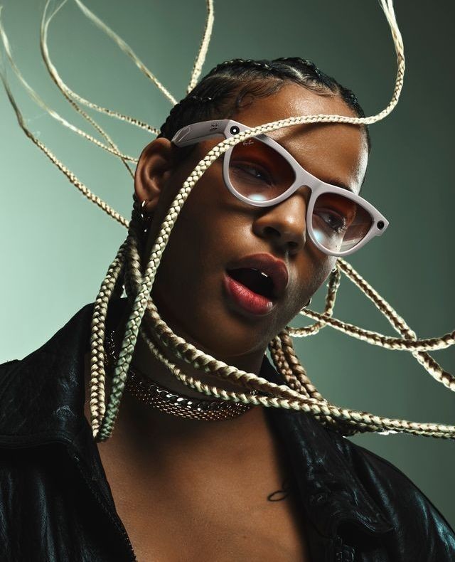 Close-up portrait of an adult woman wearing sunglasses and a black jacket, showcasing blonde braids against a muted green background.