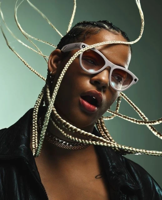 Close-up portrait of an adult woman wearing sunglasses and a black jacket, showcasing blonde braids against a muted green background.