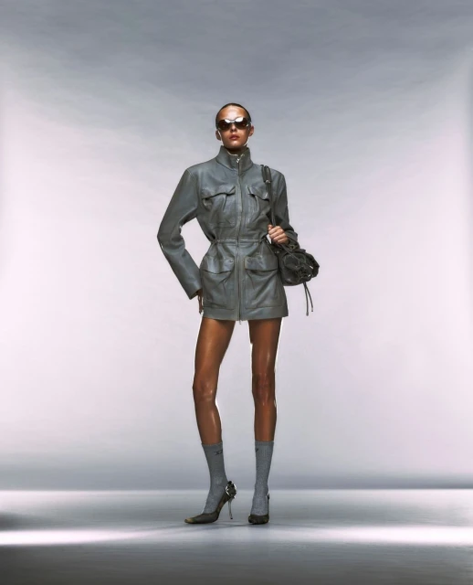 Fashion shot of a woman posing in a grey jacket dress, matching heels, sunglasses, and a dark handbag against a white backdrop.