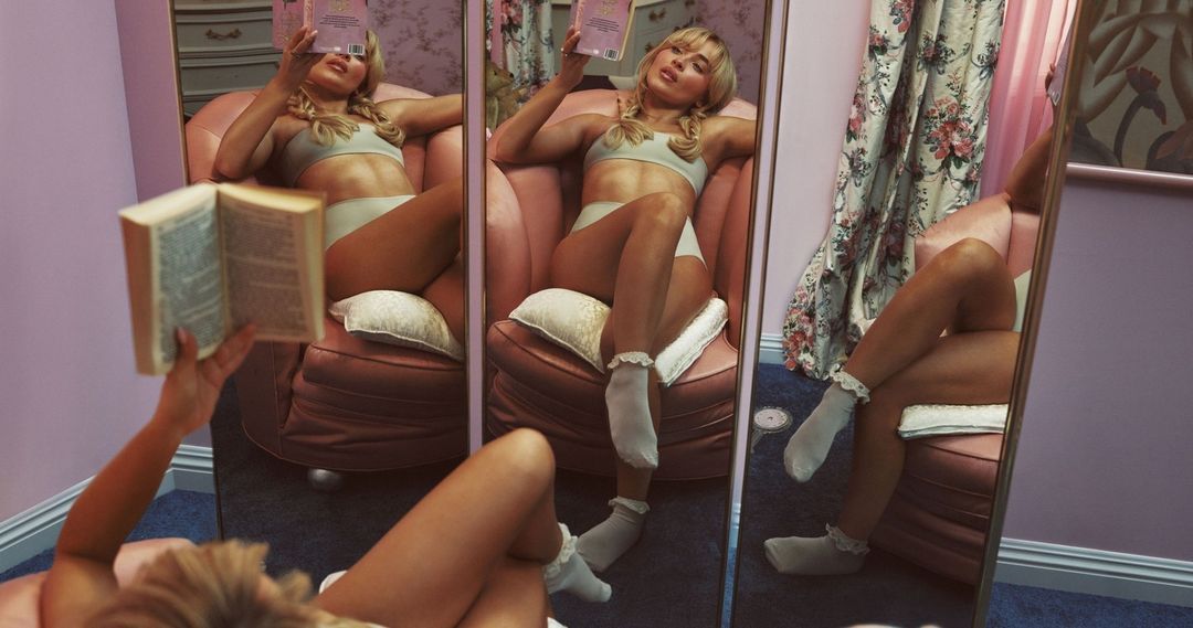 Young woman in underwear and socks sits in a chair reading a book, reflected in mirrors in a pink-toned interior.