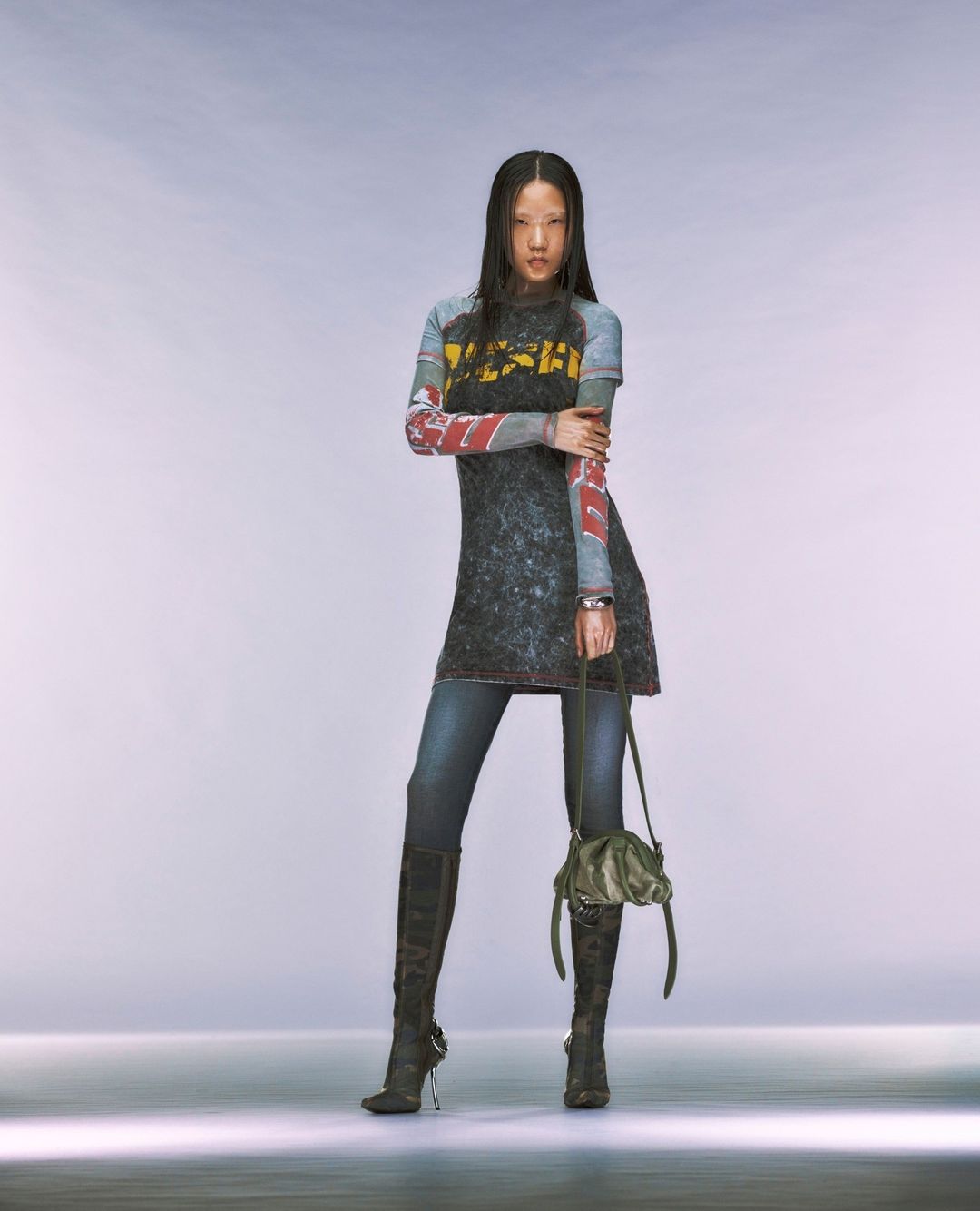 Full-length shot of a woman with dark hair posing in a black graphic top, denim jeans, and black high-heeled boots in a studio setting.