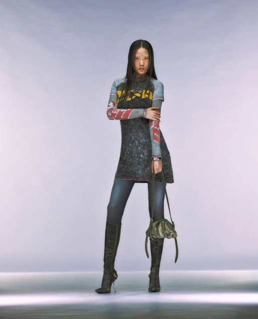 Full-length shot of a woman with dark hair posing in a black graphic top, denim jeans, and black high-heeled boots in a studio setting.