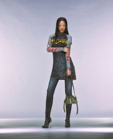 Full-length shot of a woman with dark hair posing in a black graphic top, denim jeans, and black high-heeled boots in a studio setting.