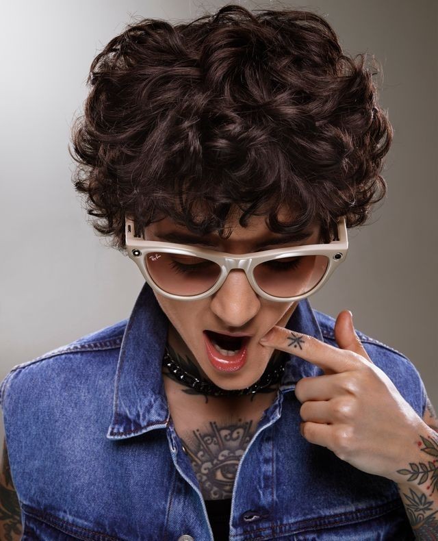 Stylish portrait of a person with curly hair, tattoos, and sunglasses wearing a denim vest against a neutral grey studio background.