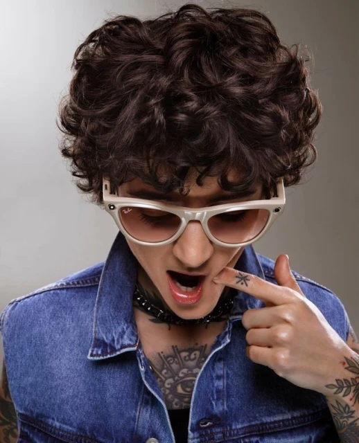 Stylish portrait of a person with curly hair, tattoos, and sunglasses wearing a denim vest against a neutral grey studio background.