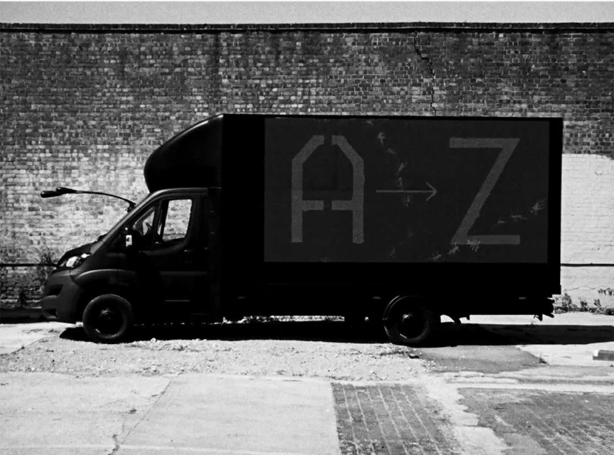 Black delivery truck with the text A to Z parked on a concrete lot in front of a brick wall in a black and white photograph.