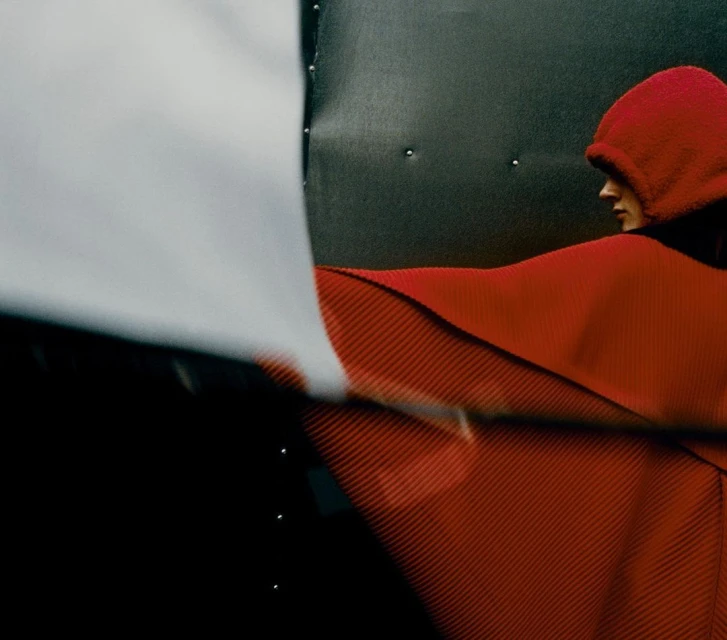 Side view of a person in a red hooded garment with a pleated texture, juxtaposed against a backdrop of black and white panels.