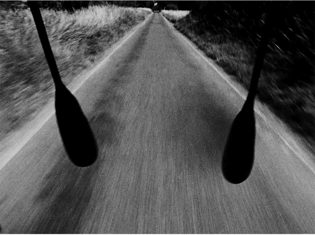 Black and white road perspective with blurred motion and silhouettes of oars, bordered by grassy fields under natural lighting.