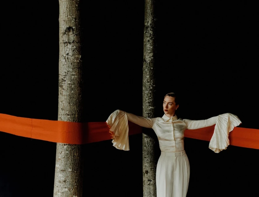 Elegant woman in white attire poses between two trees with an orange sash, set against a stark black backdrop in a studio.