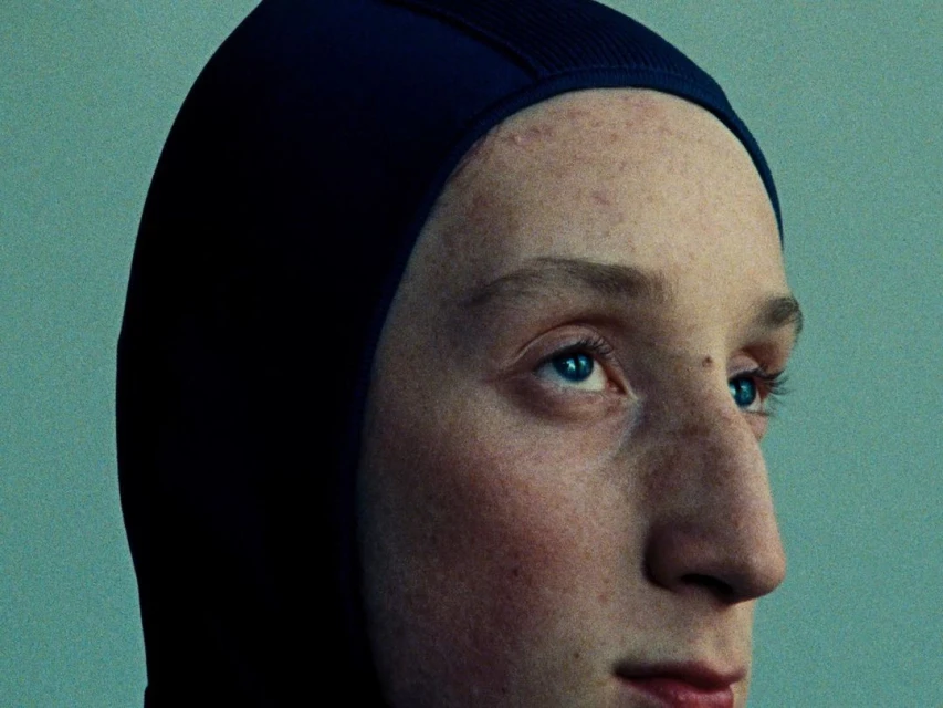 Close-up portrait of a person with blue eyes and freckles wearing a dark blue swim cap, against a teal-colored wall backdrop.