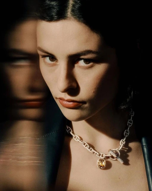Close-up portrait of a young adult female wearing a chain necklace with a yellow gemstone pendant in a dark, moody setting.
