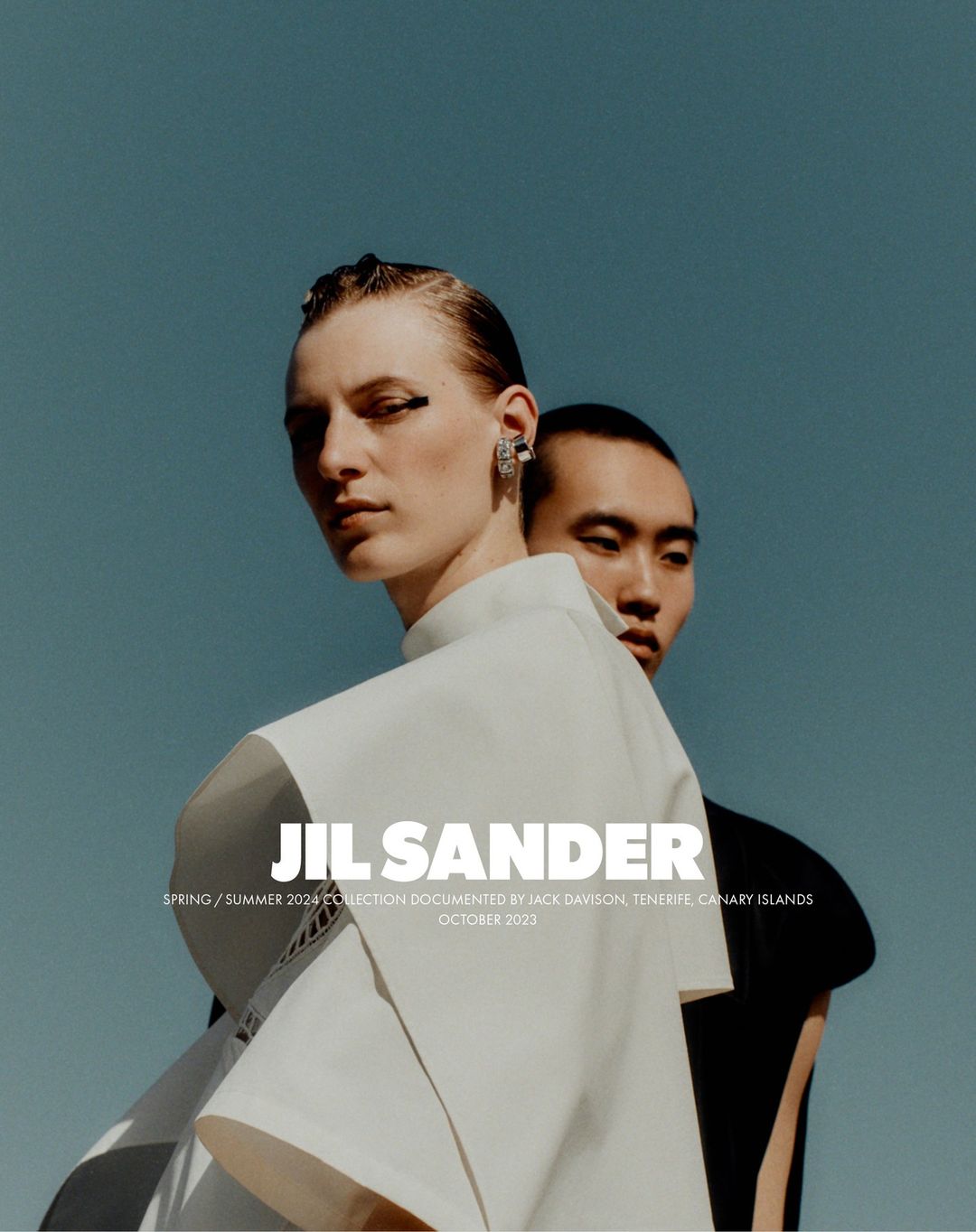 Fashion shot of two models, one fair-skinned wearing white and the other Asian, set against a blue sky. The focus is on stylish clothing.