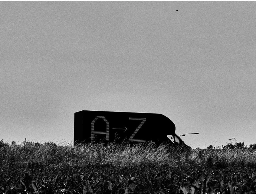 Black-and-white photo of a delivery van with 'A→Z' on its side, parked in a grassy field under a bright sky, minimalist composition.