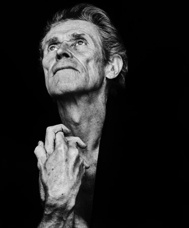 Black-and-white portrait of an elderly man looking up, his hands clasped. The image is a close-up with the background completely black.