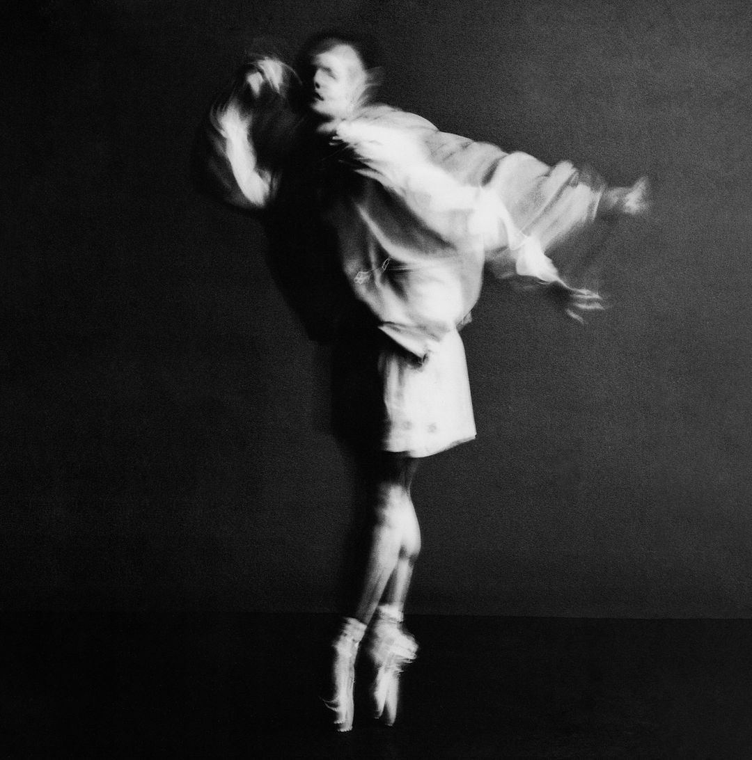 Artistic black and white dance photography: A ballet dancer in mid-air, wearing pointe shoes, with a blurred motion effect against a dark background.