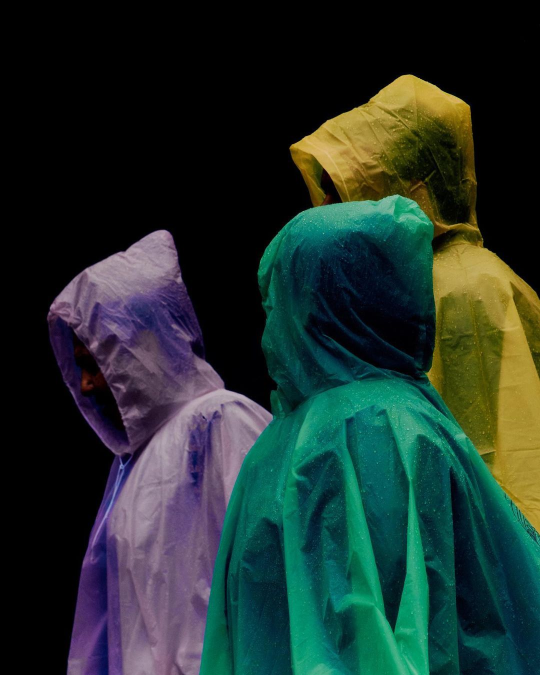 Three people in colorful translucent rain ponchos are closely grouped against a stark black background, creating a vibrant, abstract composition.