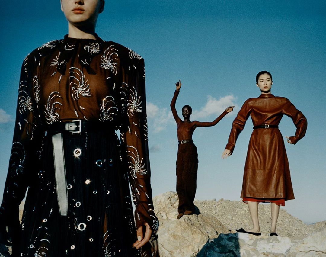 Three fashion models pose outdoors on a rocky surface against a blue sky. Two of the models are wearing long dresses.