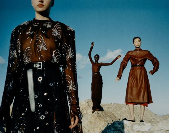 Three fashion models pose outdoors on a rocky surface against a blue sky. Two of the models are wearing long dresses.