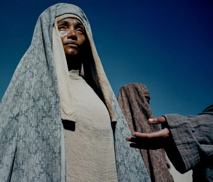 Three people outdoors, wearing traditional draped clothing, one looking up at the sky with blue sky visible in the background.