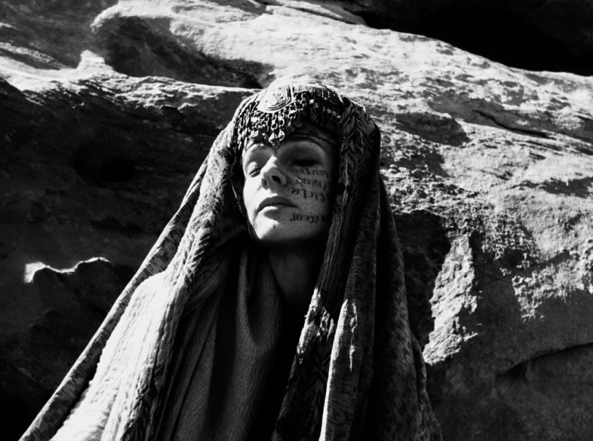 A monochrome image features a person in desert-like setting, adorned with face markings and a headdress, complemented by rock formations.