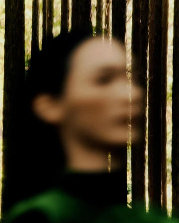 Woman portrait with blurred face, stands behind silhouetted trees in a forest-like setting. Soft focus aesthetic adds a dreamy quality.