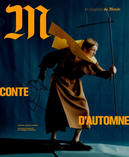 Fashion magazine cover featuring a model in a brown outfit with a yellow collar, holding a rod and wires, against a blue background.