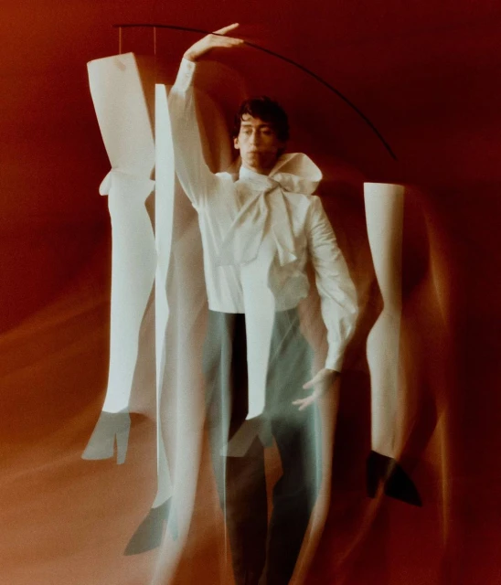 Man in motion posing with white sculptural legs against a red background, wearing a tied white shirt, and gray pants. High-fashion studio shot.