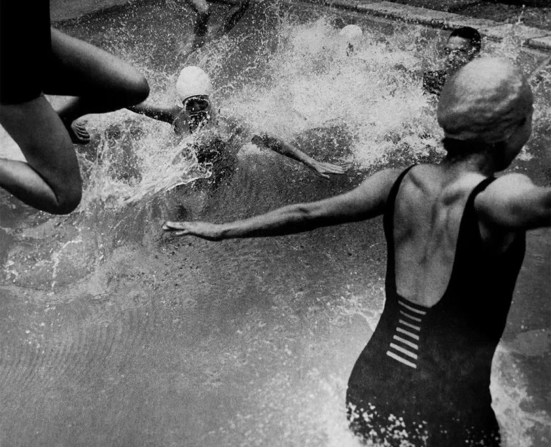 Monochrome photograph of several people swimming in a pool, creating splashes and motion in the water, wearing swimsuits and caps.