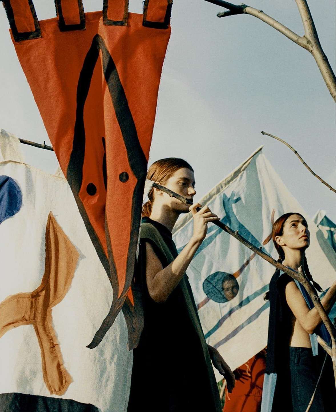 Two women stand behind painted flags in an outdoor environment, posing as though waiting for a signal, under a clear sky during the day.