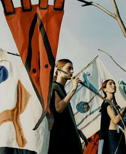 Two women stand behind painted flags in an outdoor environment, posing as though waiting for a signal, under a clear sky during the day.