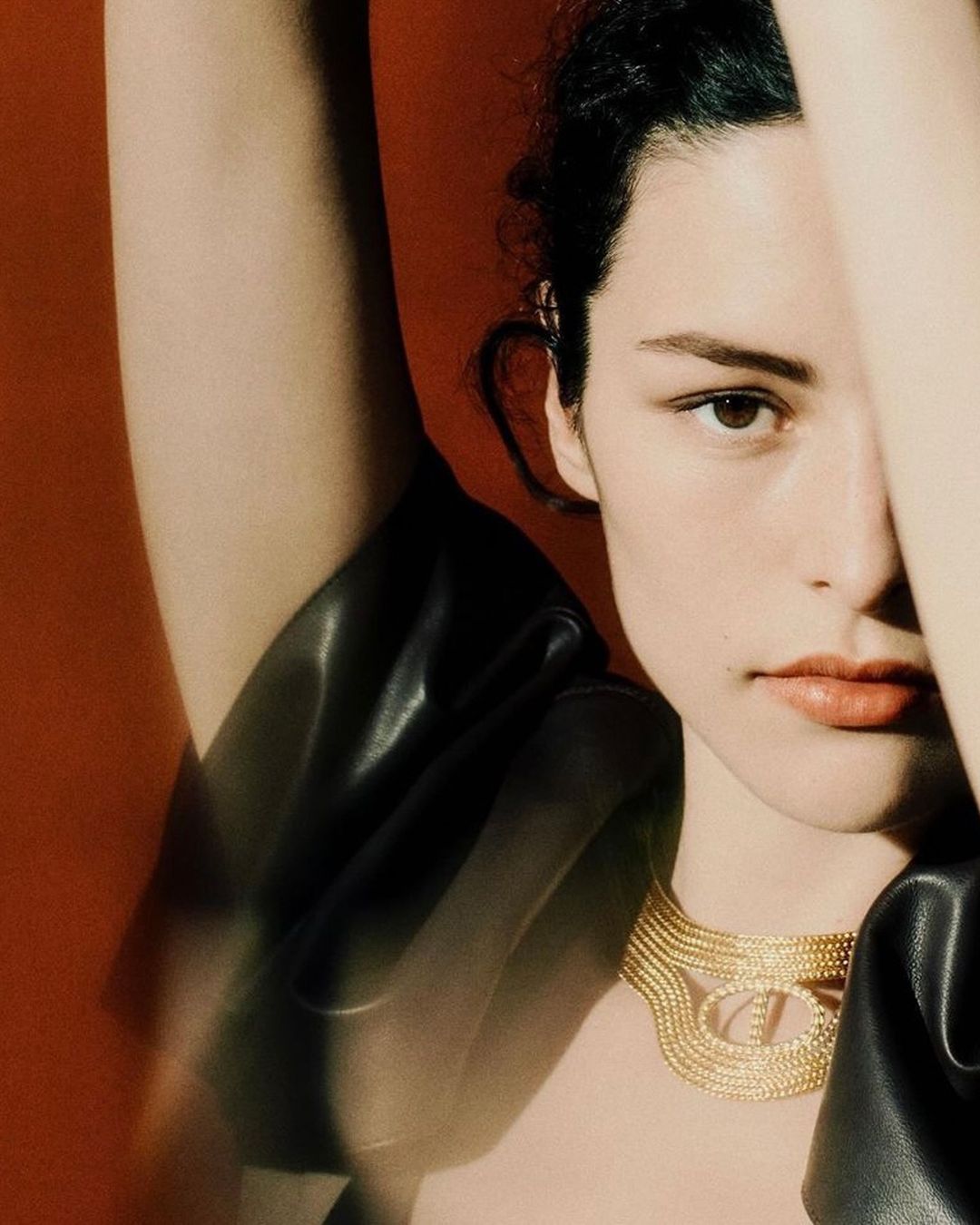 Close-up portrait of a young woman with fair skin and dark hair, wearing a gold necklace against a reddish-brown background in a studio.
