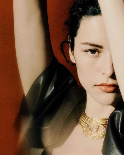Close-up portrait of a young woman with fair skin and dark hair, wearing a gold necklace against a reddish-brown background in a studio.
