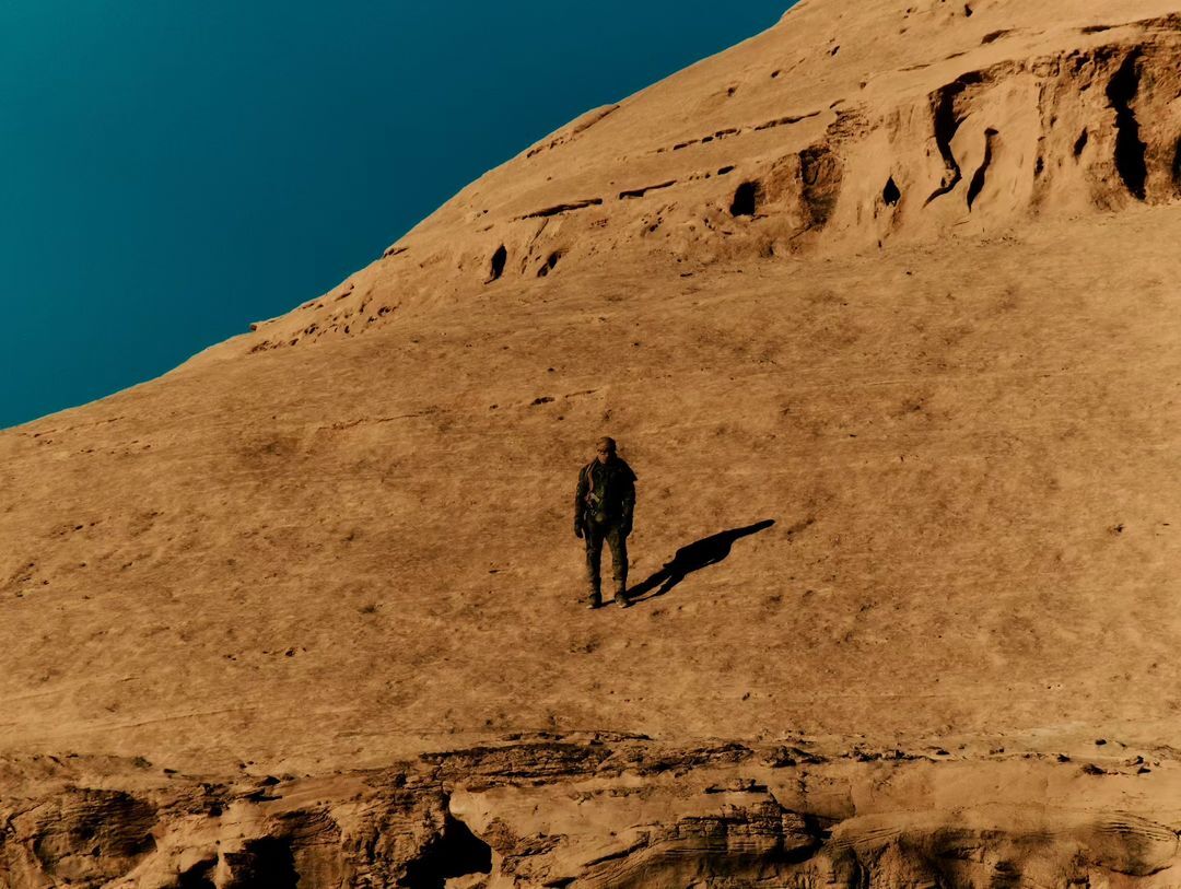 A person stands in a desert landscape next to a large rock formation under a clear blue sky, casting a long shadow in the sunlight.