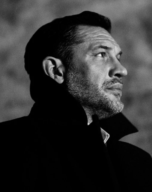 Black and white portrait of an adult man with short hair and a beard wearing a dark overcoat in an indoor setting.