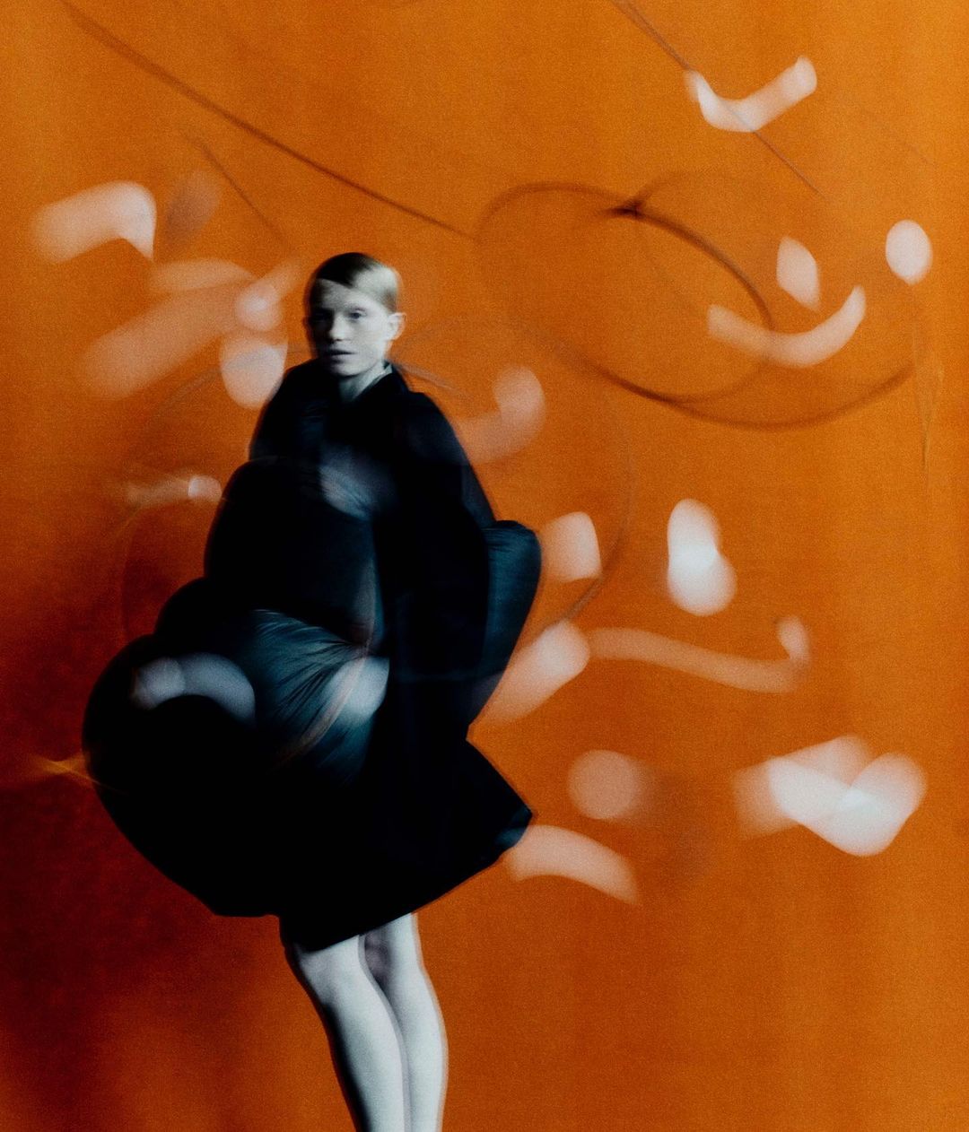 Fashion shoot of an adult woman in a black dress and white tights standing in front of an orange background with blurred, white patterns.