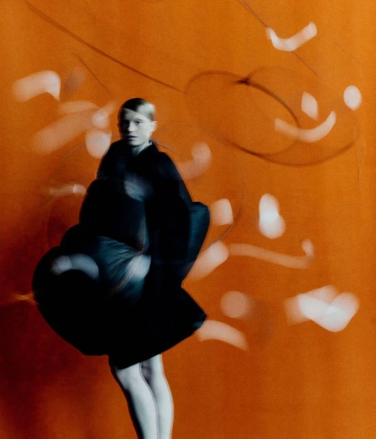 Fashion shoot of an adult woman in a black dress and white tights standing in front of an orange background with blurred, white patterns.