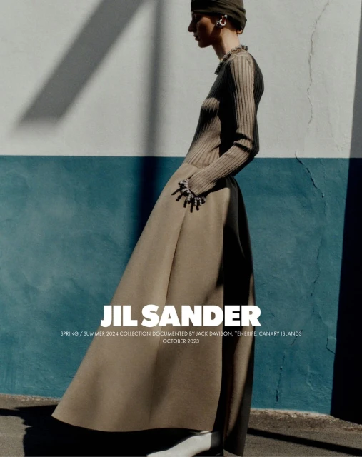 Fashion editorial photo featuring a woman wearing a long brown dress with statement bracelet accessories, posing against a blue and white wall.