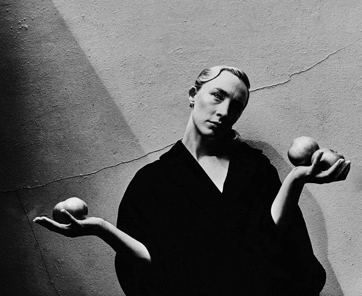 Monochrome portrait of an adult female holding two fruits in her hands in front of a grey textured wall with subtle lighting.