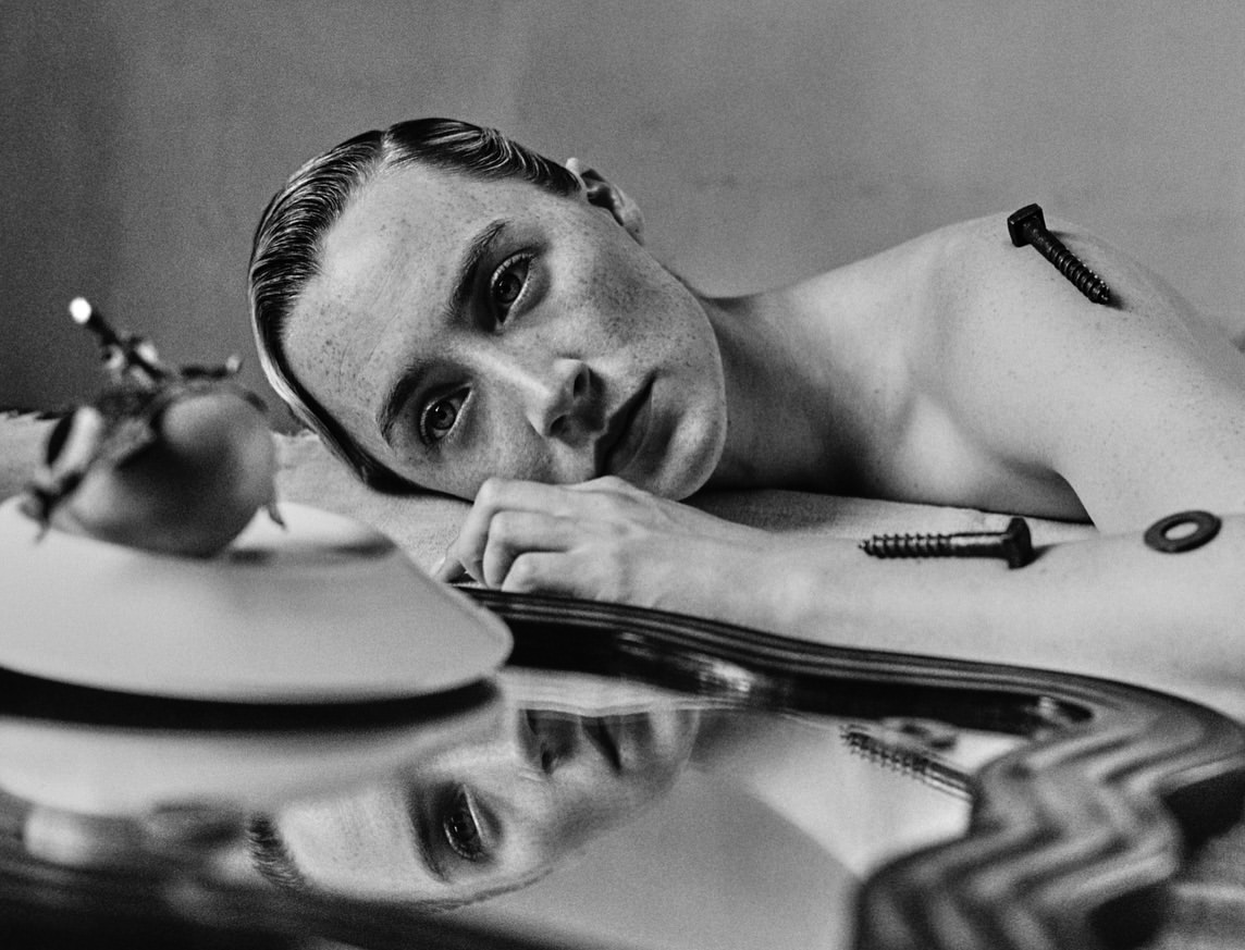 Monochrome portrait of a fair-skinned person lying down with screws and hardware on their body, with a tomato on a plate.