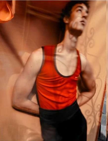 A young adult man in a red tank top and dark pants looks up in a studio environment, captured with a slightly blurred effect, indoor.